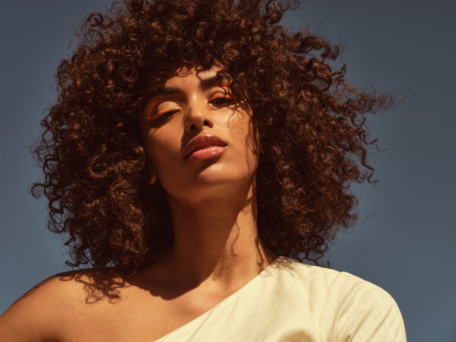 Porträt einer Frau mit lockigem, braunem Haar, orangefarbenem Eyeliner und glänzenden Lippen vor einem blauen Himmel.