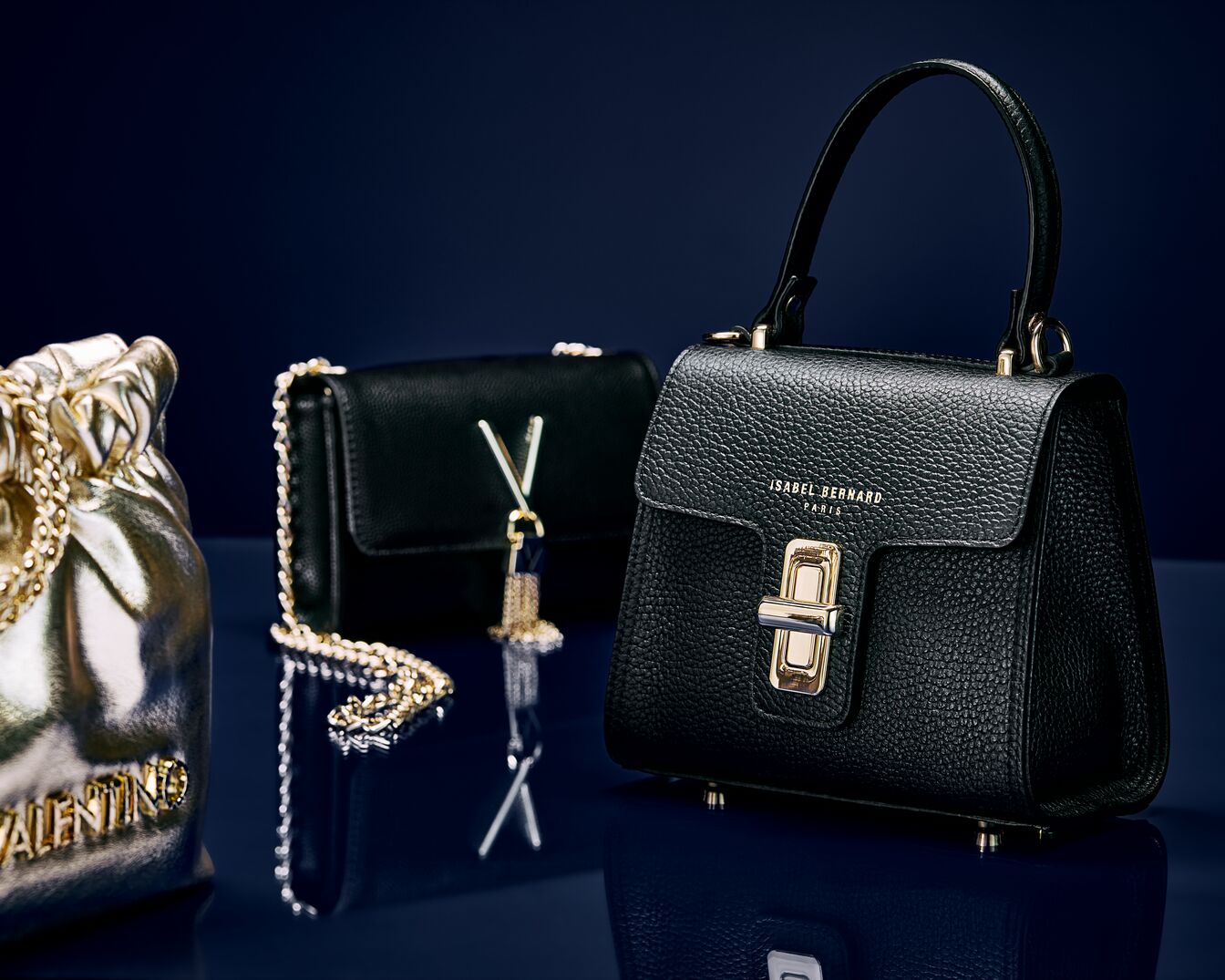 Na ciemnym tle znajdują się trzy torebki: złota torebka Valentino, czarna torebka na łańcuszku z symbolem V oraz czarna torebka Isabel Bernard z krótkim uchwytem.