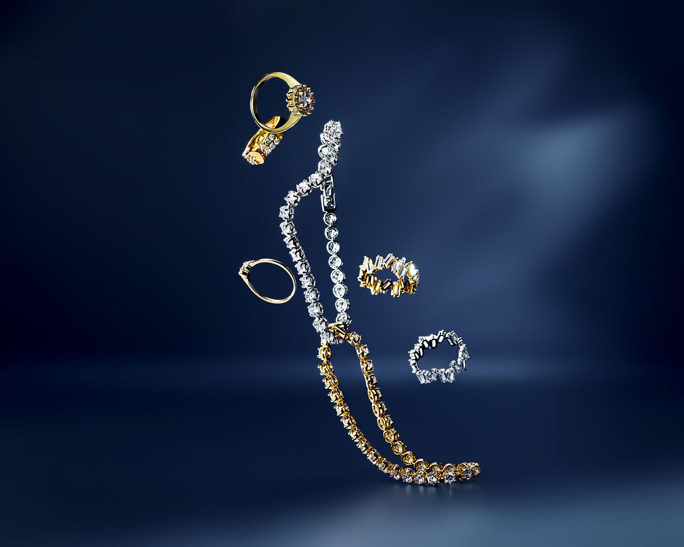 Na ciemnoniebieskim tle unoszą się złote i srebrne pierścionki oraz naszyjnik wysadzany diamentami, tworząc elegancką kompozycję.