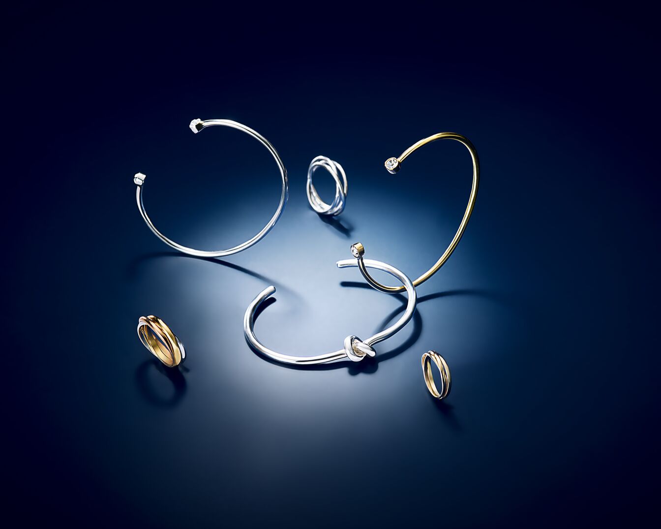 Na ciemnoniebieskim tle leżą pierścionki i bransoletki w kolorze srebrnym i złotym, tworząc elegancką kompozycję.