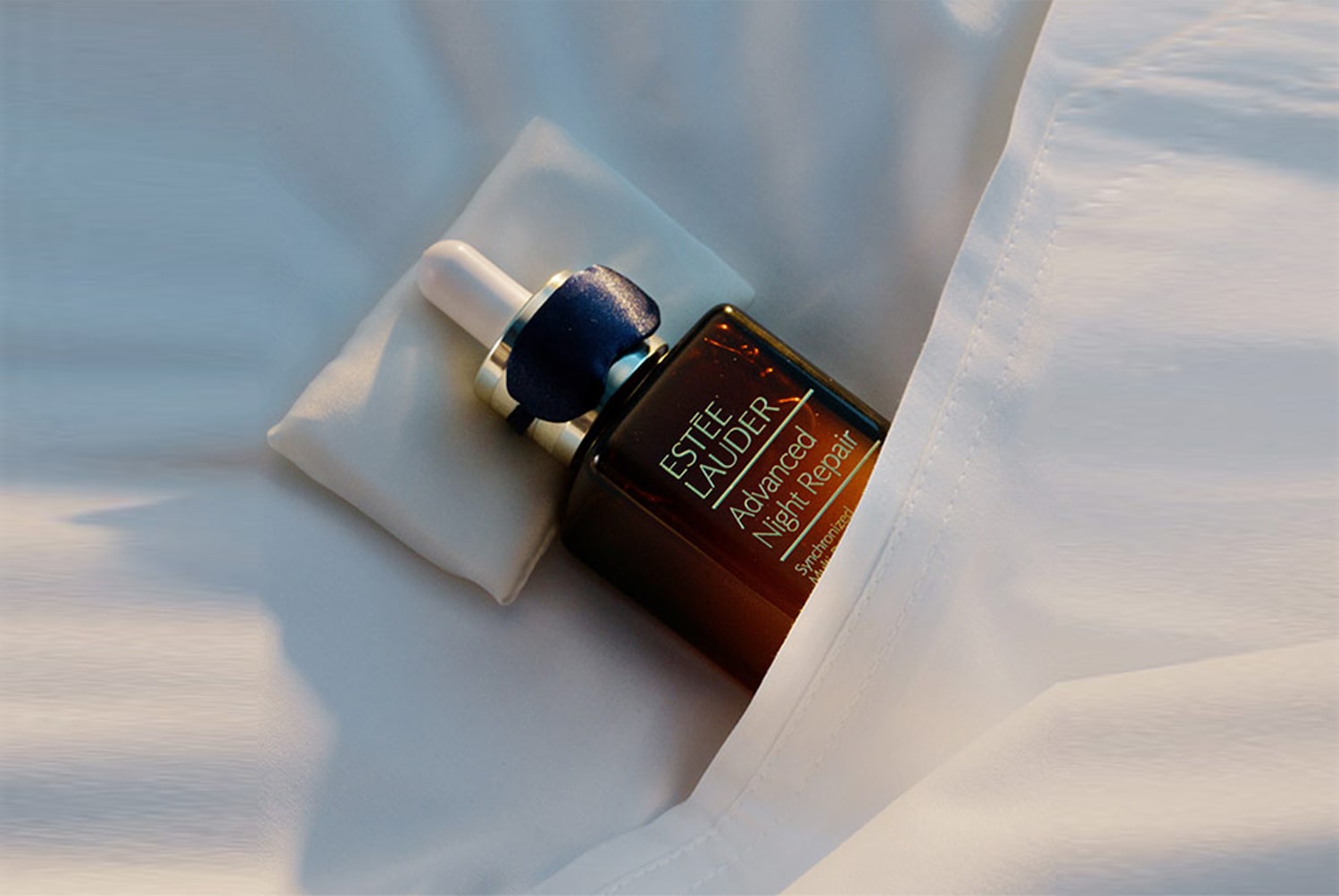 Az Estée Lauder Advanced Night Repair szérum egy barna üvegben, fehér párnán és fehér vásznon pihenve látható.