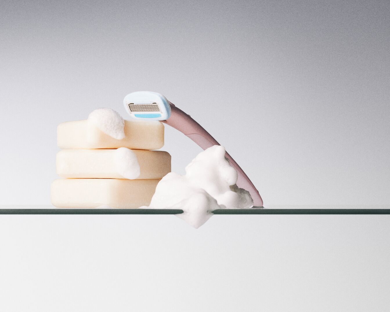 Auf einem Glasregal liegen drei cremefarbene Seifenstücke mit Schaum und ein pinkfarbener Rasierer mit hellblauem Klingenkopf.