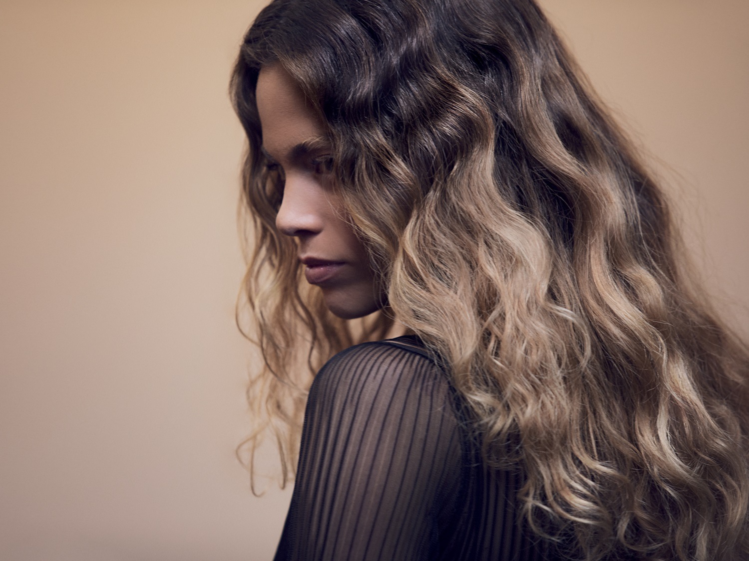 Kobieta z długimi, falowanymi włosami w odcieniach brązu i blondu, ubrana w czarną, prześwitującą bluzkę, spogląda w dół.