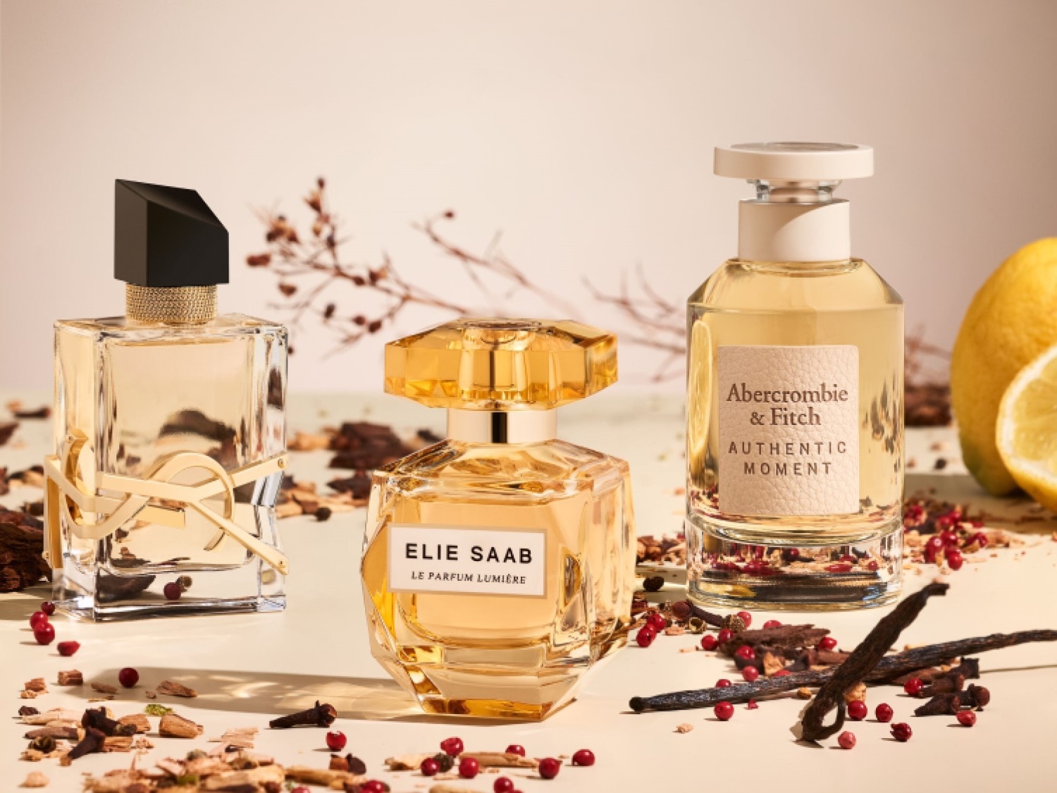 Tri zlatisté parfumy od YSL, Elie Saab a Abercrombie & Fitch stoja na svetlom pozadí, posypané drobnými kúskami dreva, červenými bobuľami a kúskami vanilky.
