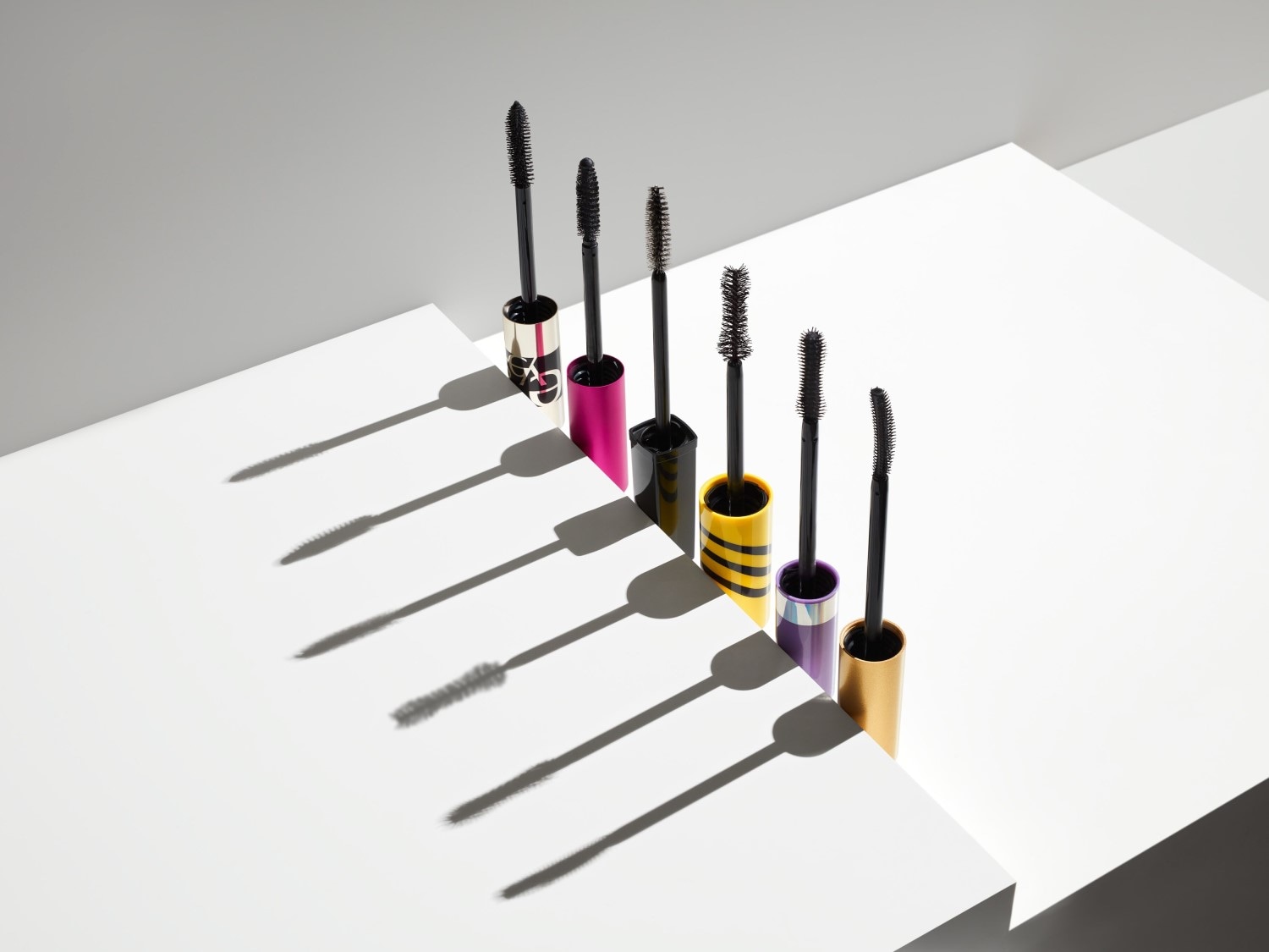 Eine Reihe von Mascaras in verschiedenen Farben, darunter Gold, Pink, Gelb und Lila, stehen auf einer weißen Oberfläche und werfen lange Schatten.