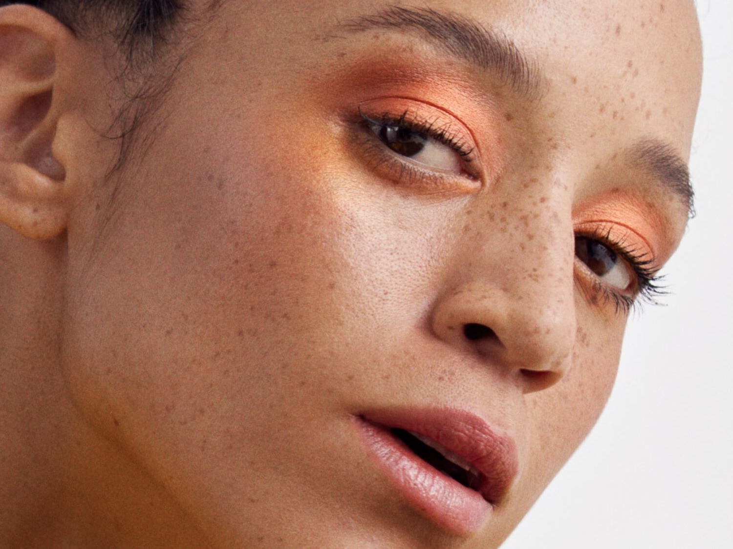 Nahaufnahme eines Frauengesichts mit Sommersprossen und einem auffälligen orangefarbenen Lidschatten, der den Blick betont.