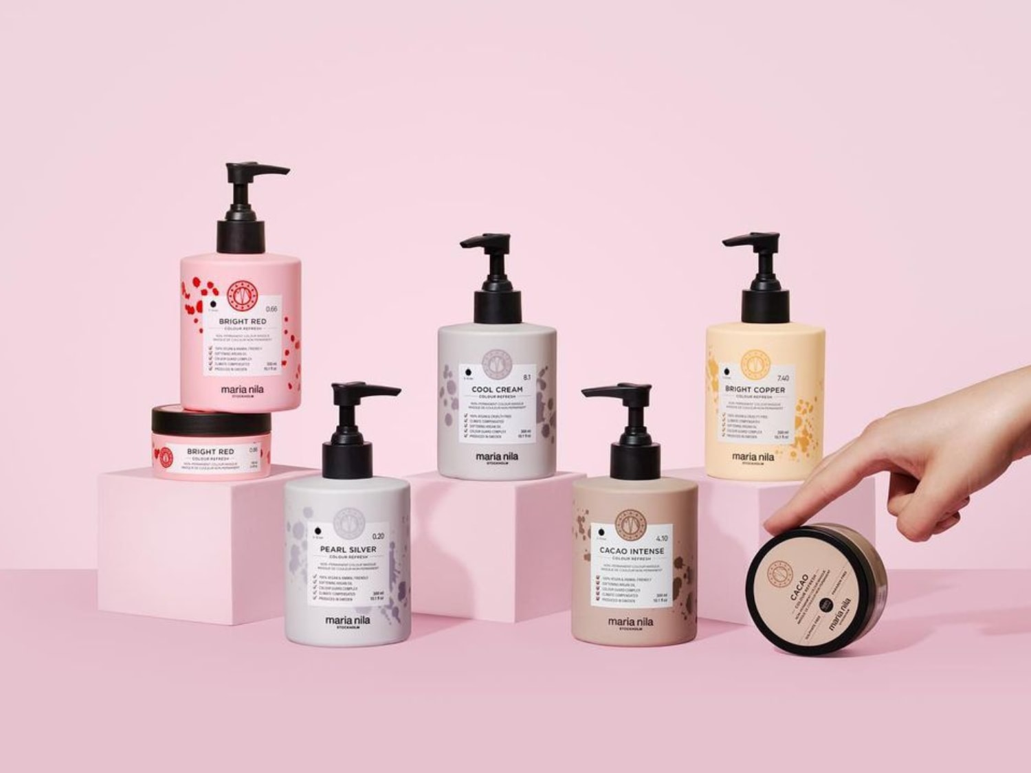 Eine Auswahl an Maria Nila Farbpflegeprodukten in verschiedenen Farbtönen, darunter Bright Red, Cool Cream, Pearl Silver, Bright Copper und Cacao Intense, präsentiert auf rosa Podesten vor einem passenden Hintergrund.