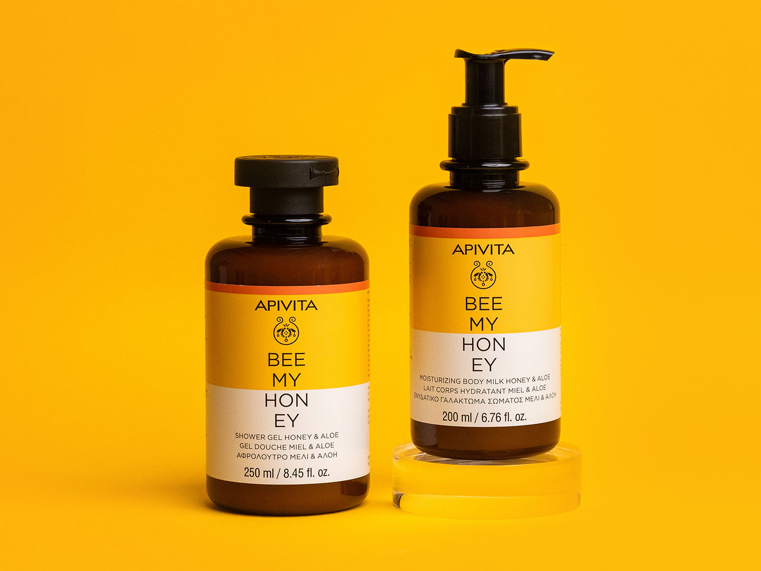 Dos botellas de Apivita Bee My Honey, una de gel de ducha y otra de leche corporal hidratante con miel y aloe, sobre un fondo amarillo.