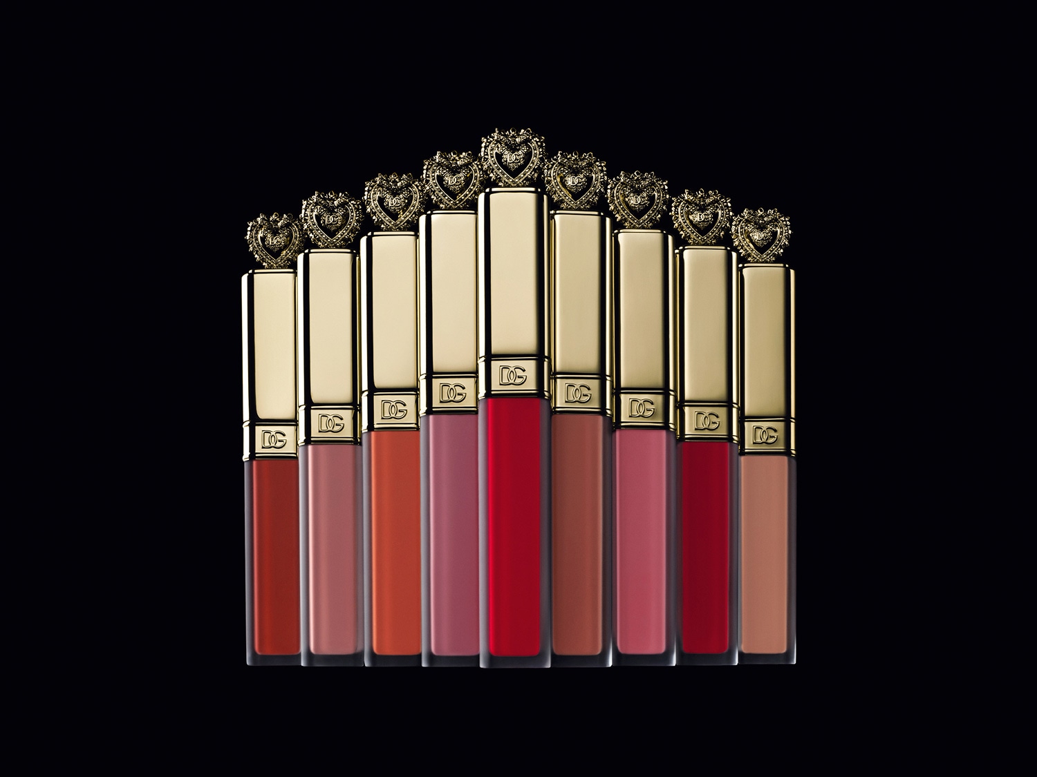 Aquí tienes una colección de labiales Dolce & Gabbana en varios tonos, cada uno con un tapón dorado adornado con un corazón enjoyado, sobre un elegante fondo negro.