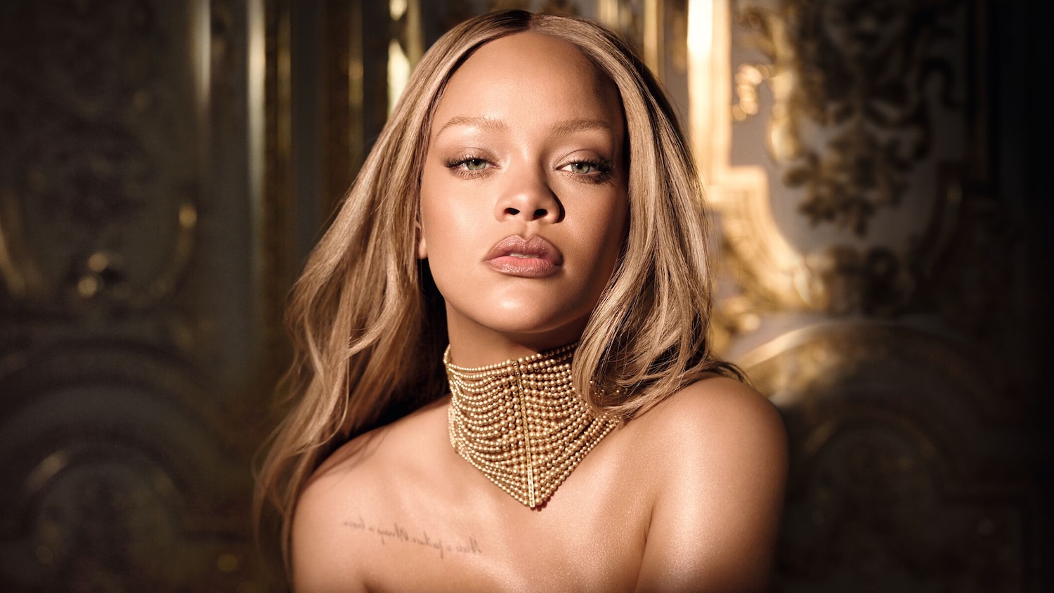 Gros plan sur Rihanna, les cheveux blond miel, portant un collier doré complexe qui met en valeur son teint éclatant et son regard perçant.