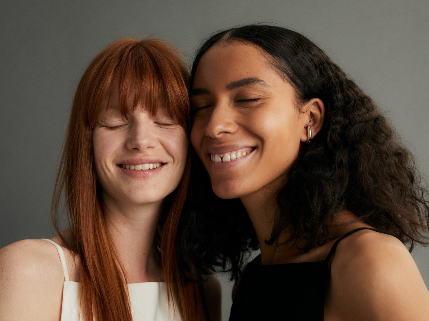 Zwei strahlende Frauen, eine mit roten, glatten Haaren und die andere mit dunklen, lockigen Haaren, lächeln mit geschlossenen Augen und zeigen ihre strahlenden Zähne.