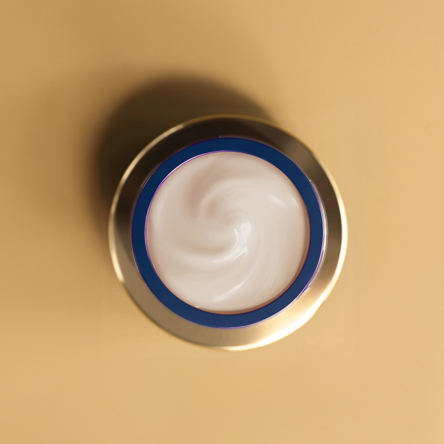 Nahaufnahme einer goldenen Dose mit blauem Rand, gefüllt mit einer weißen Creme mit gewirbelter Textur, die auf einem beigen Hintergrund steht.