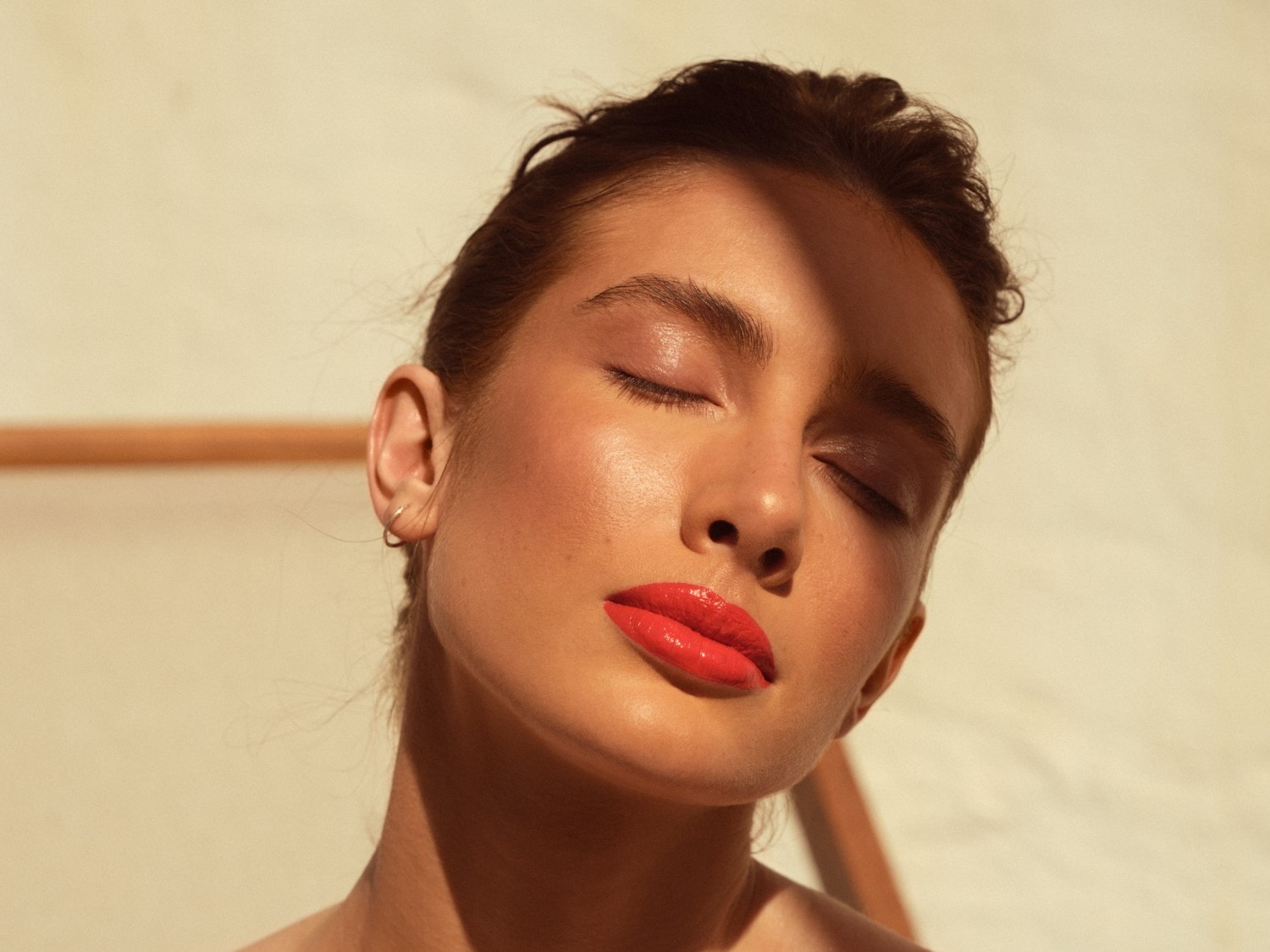 Une femme aux yeux clos, illuminée par le soleil, arbore des lèvres rouge corail brillantes, capturant un instant de beauté sereine.