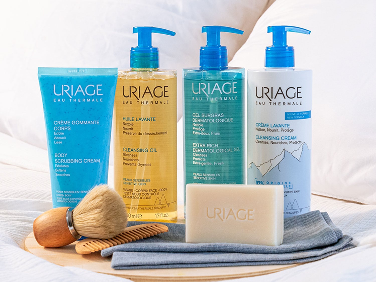 Descubre la gama Uriage Eau Thermale con crema exfoliante azul, aceite limpiador dorado, gel dermatológico y crema limpiadora en envases con dosificador azul, junto a un jabón, brocha y peine sobre una toalla.