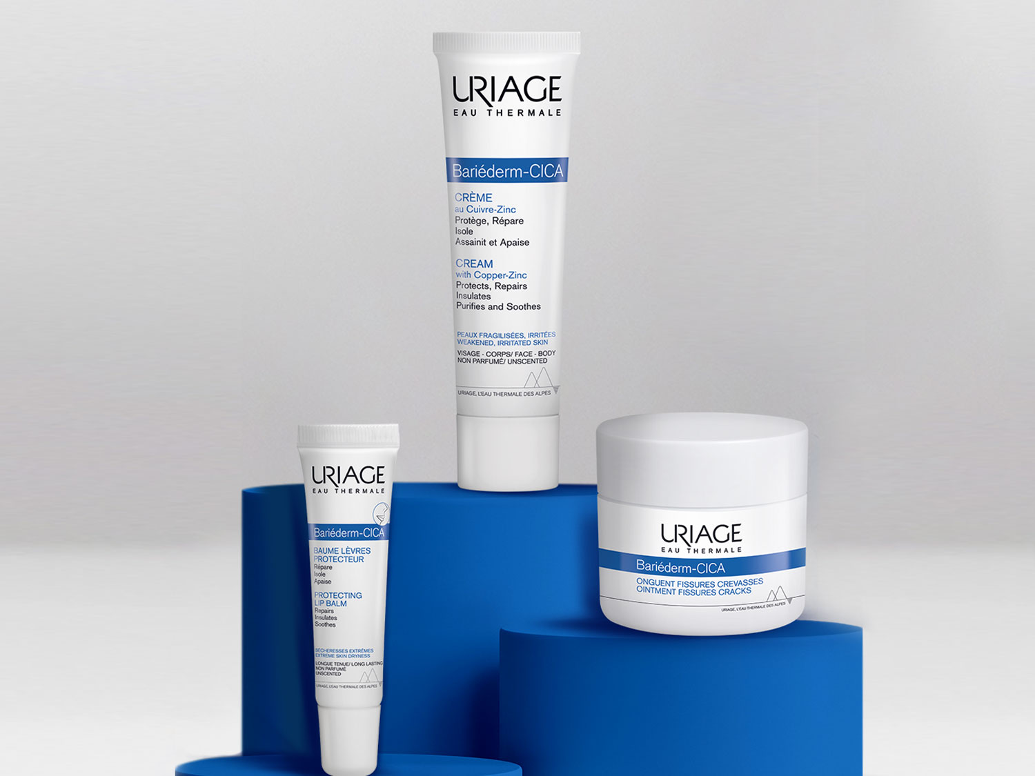 Aquí hay un conjunto de productos Uriage Bariéderm-CICA, que incluyen crema, bálsamo labial protector y ungüento para fisuras y grietas, todos en envases blancos con detalles azules.