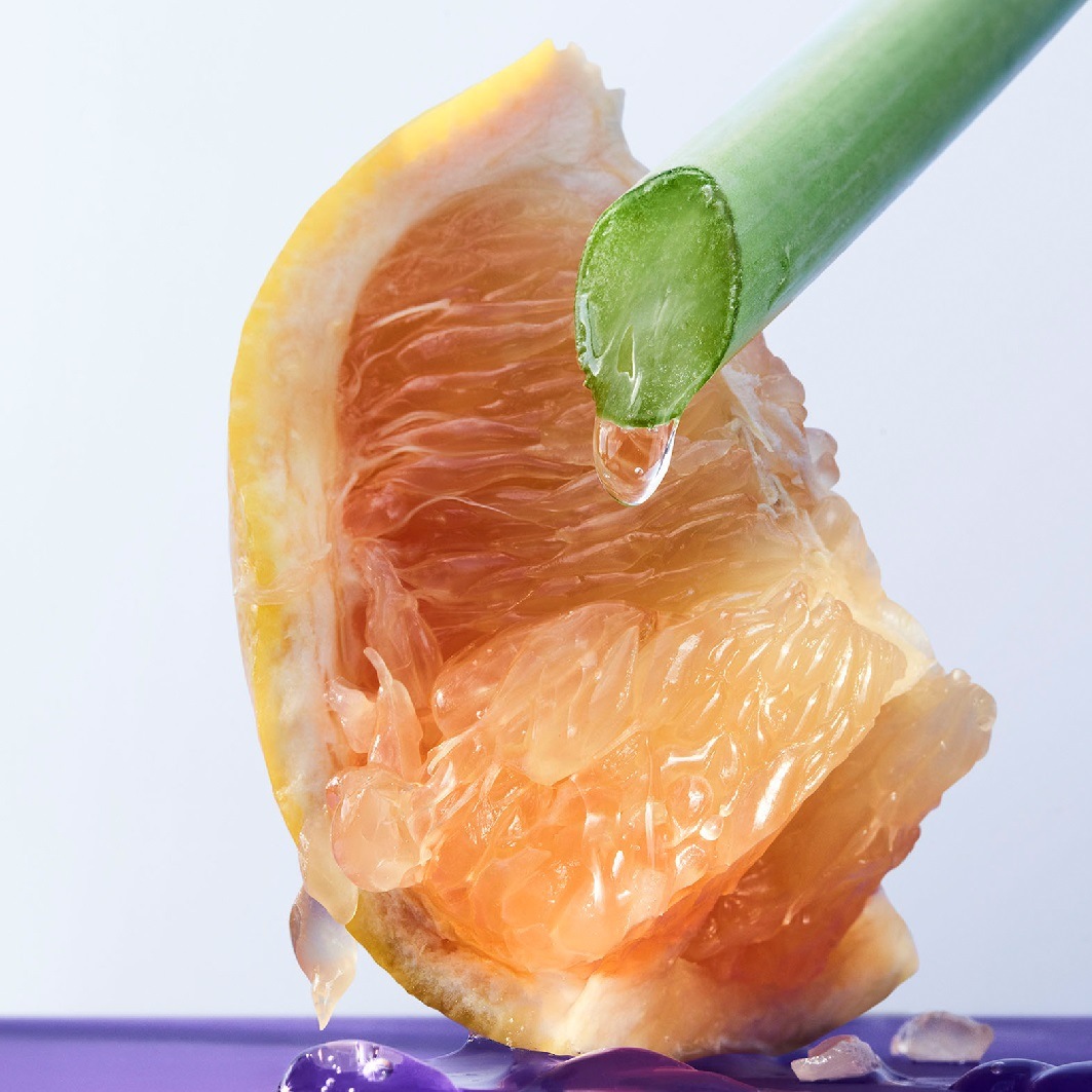 Ein saftiger Grapefruit-Schnitz mit einem grünen Stiel, von dem ein klarer Tropfen auf eine violette Oberfläche fällt.