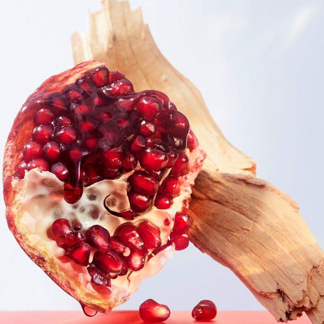 Nahaufnahme einer halben Granatapfelfrucht mit roten Samen und Saft, die neben einem Stück Holz auf einer roten Oberfläche liegt.