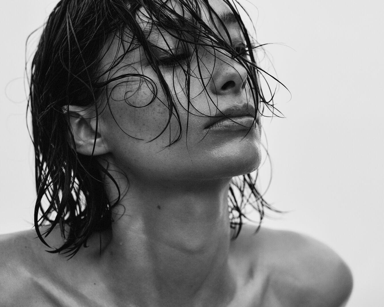 Primer plano en blanco y negro de una mujer con el pelo mojado peinado hacia la cara y los ojos cerrados, mostrando una piel fresca y húmeda.