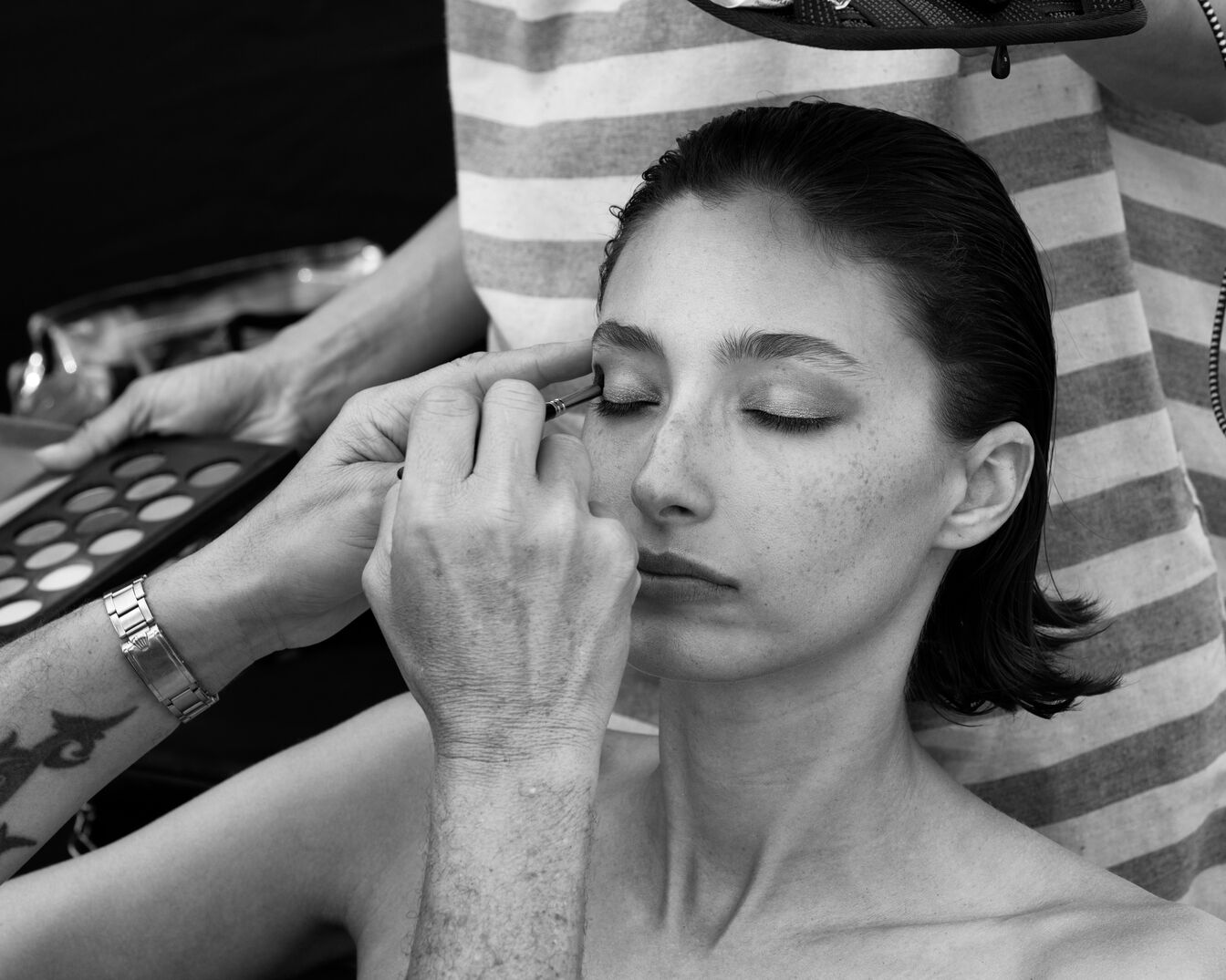 En la imagen en blanco y negro, una maquilladora aplica delineador de ojos a una modelo de piel clara con pecas y cabello oscuro peinado hacia atrás, usando una paleta de sombras de ojos.