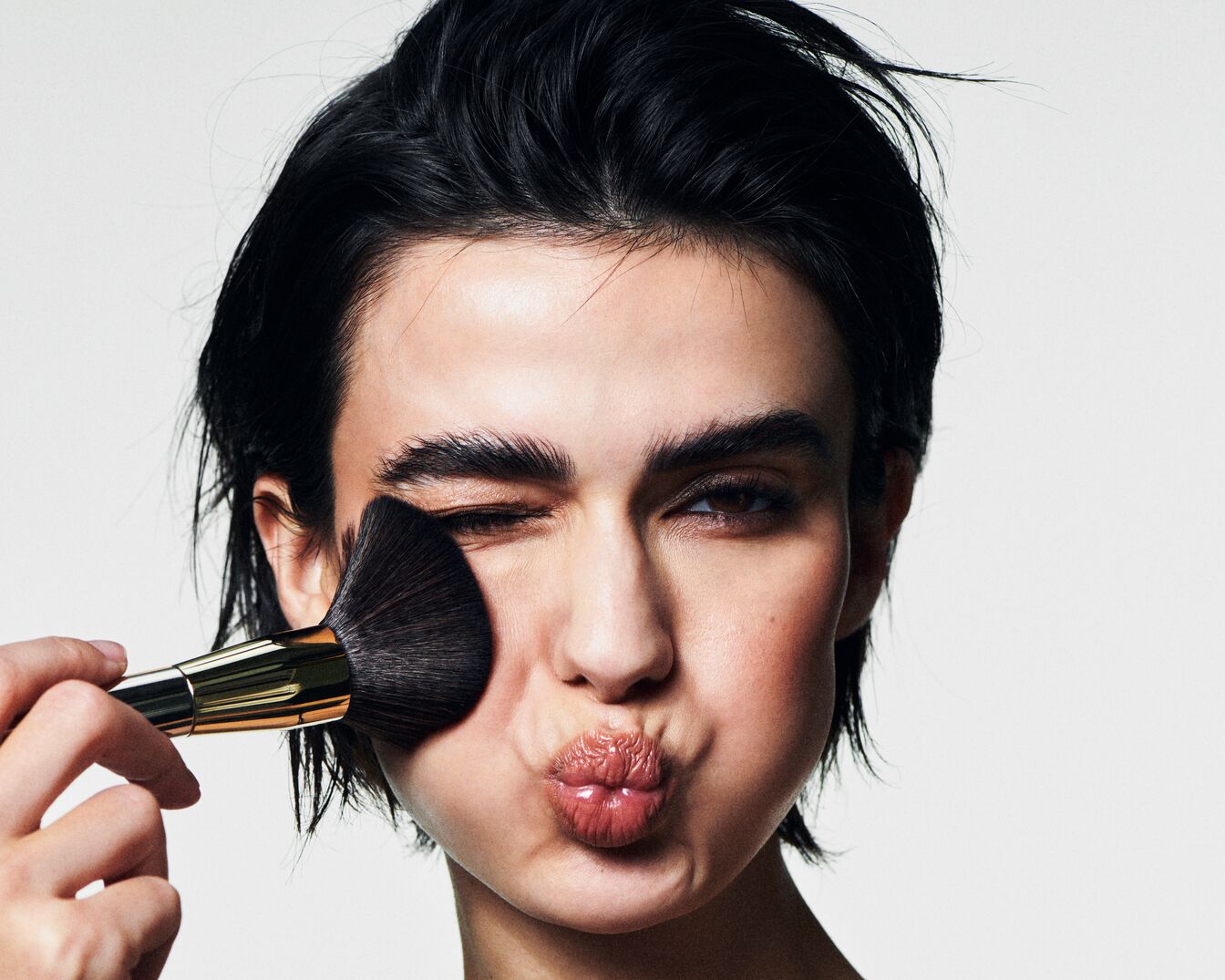 Nahaufnahme einer Frau, die mit einem großen Pinsel Make-up aufträgt, während sie die Lippen spitzt.