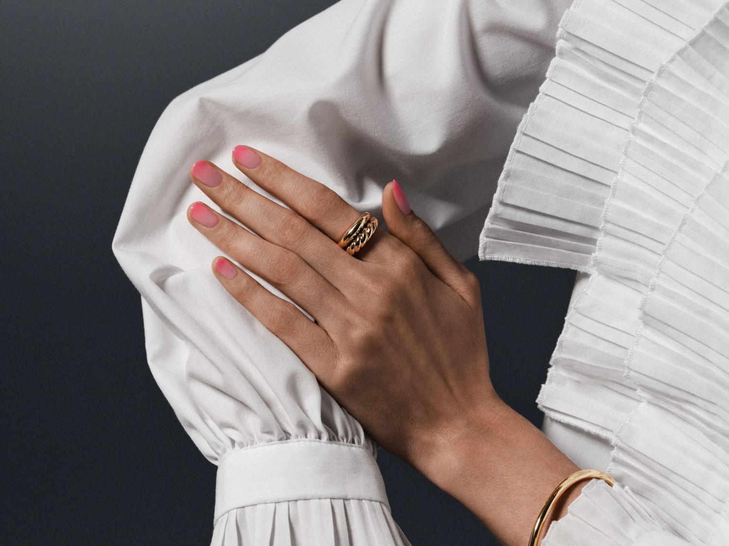 Main tenant un chemisier blanc plissé, une bague dorée à plusieurs rangs orne son doigt et un bracelet doré son poignet, tandis que ses ongles affichent un dégradé rose.