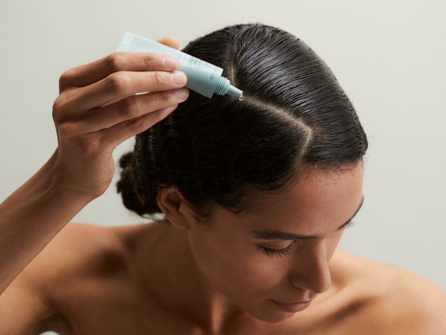 Une femme applique un sérum capillaire à partir d'un tube bleu clair sur ses cheveux mouillés et coiffés en arrière, en se concentrant sur la raie.