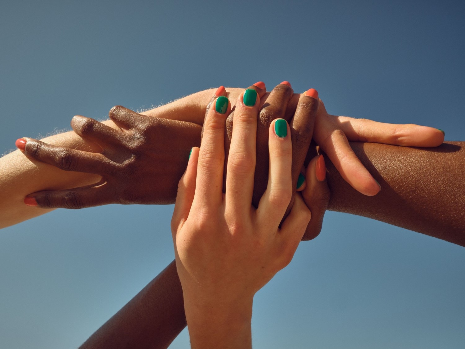 Plusieurs mains de différentes couleurs de peau sont entrelacées, certaines avec du vernis à ongles vert et d'autres avec du vernis à ongles orange, sur un fond bleu uni.