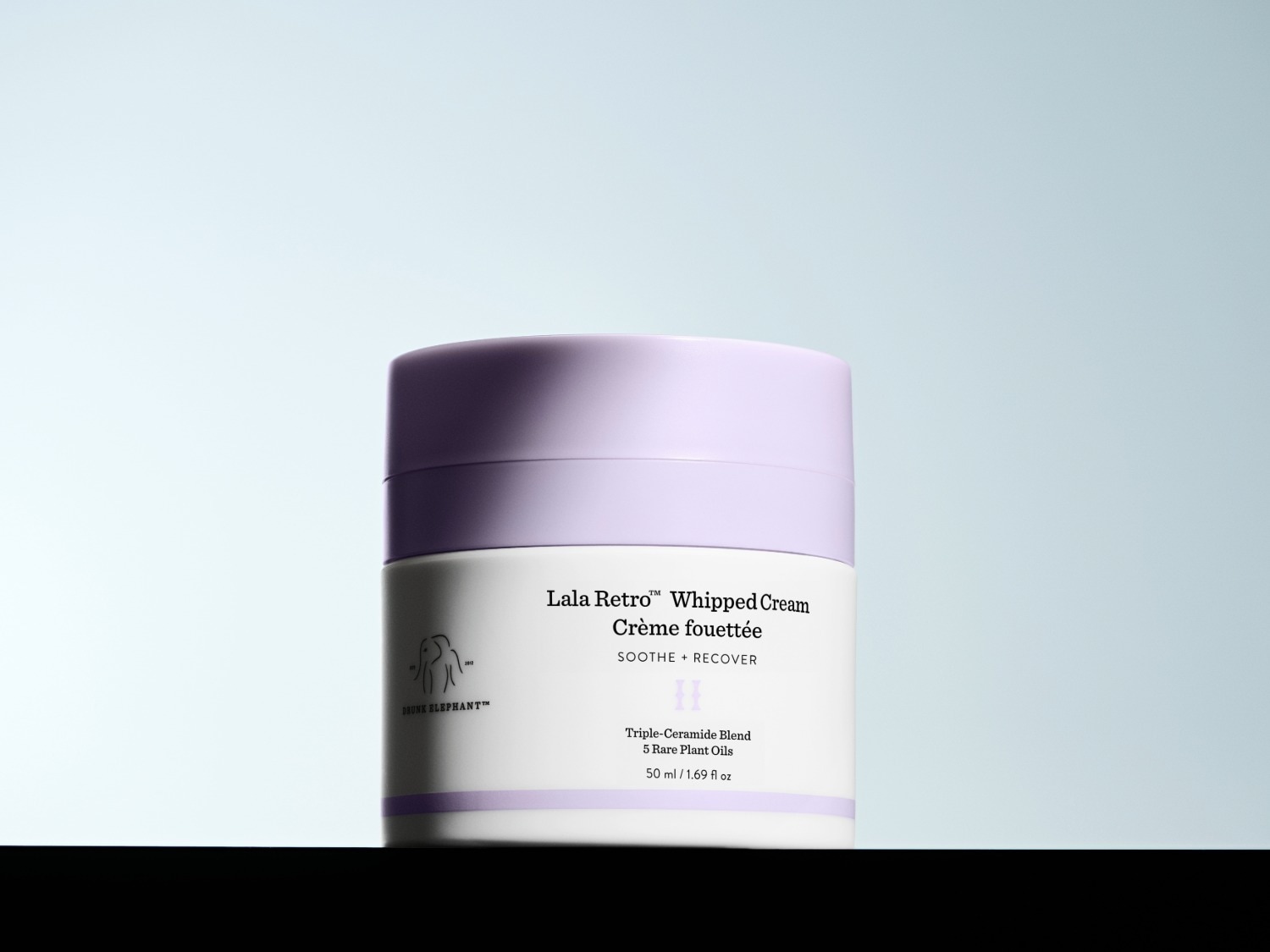 Drunk Elephant Lala Retro Whipped Cream in een wit potje met een lichtpaarse deksel, met de tekst "Soothe + Recover" en "Triple-Ceramide Blend, 5 Rare Plant Oils" erop.