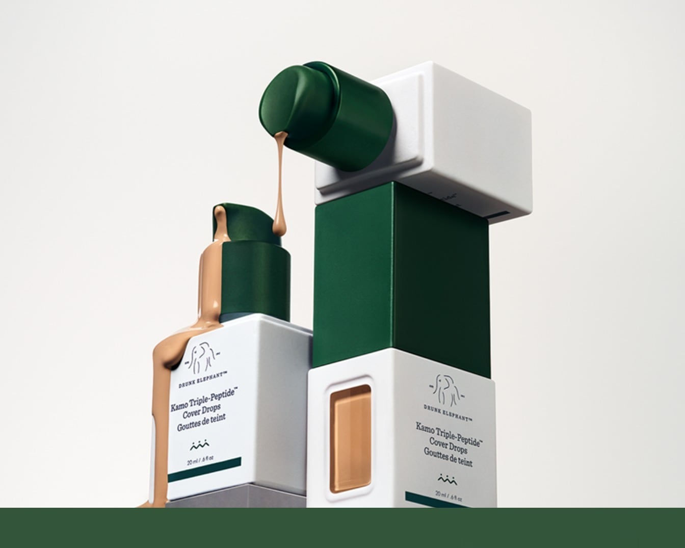 Drunk Elephant Kamo Triple-Peptide Cover Drops in een witte fles met donkergroene dop, waaruit een beige vloeistof druipt, staat naast een verpakking met een kijkvenster dat de kleur van de foundation toont.