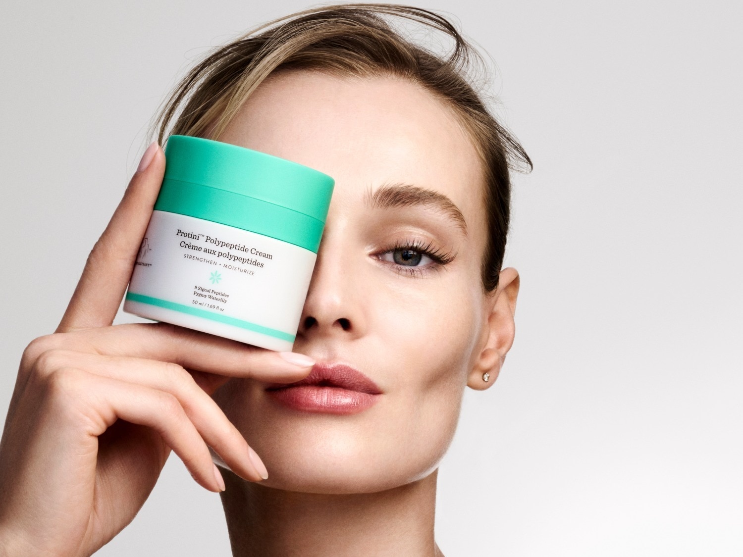 Een vrouw houdt een witte pot Protini Polypeptide Cream met een turquoise deksel voor haar gezicht, met de tekst "STRENGTHEN + MOISTURIZE" en "9 Signal Peptides Pygmy Waterlily 50 ml / 1.69 fl oz" zichtbaar.