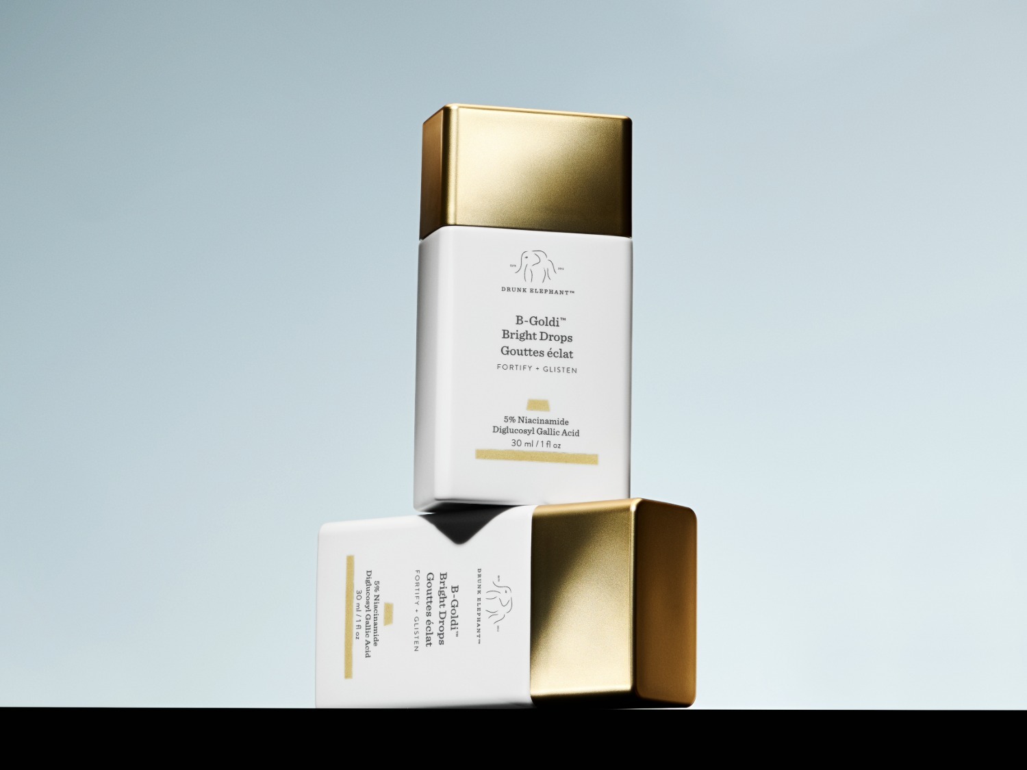 Twee witte vierkante flacons van Drunk Elephant B-Goldi Bright Drops, elk met een glanzende gouden dop, staan op elkaar gestapeld tegen een lichtblauwe achtergrond, met de productinformatie duidelijk leesbaar.