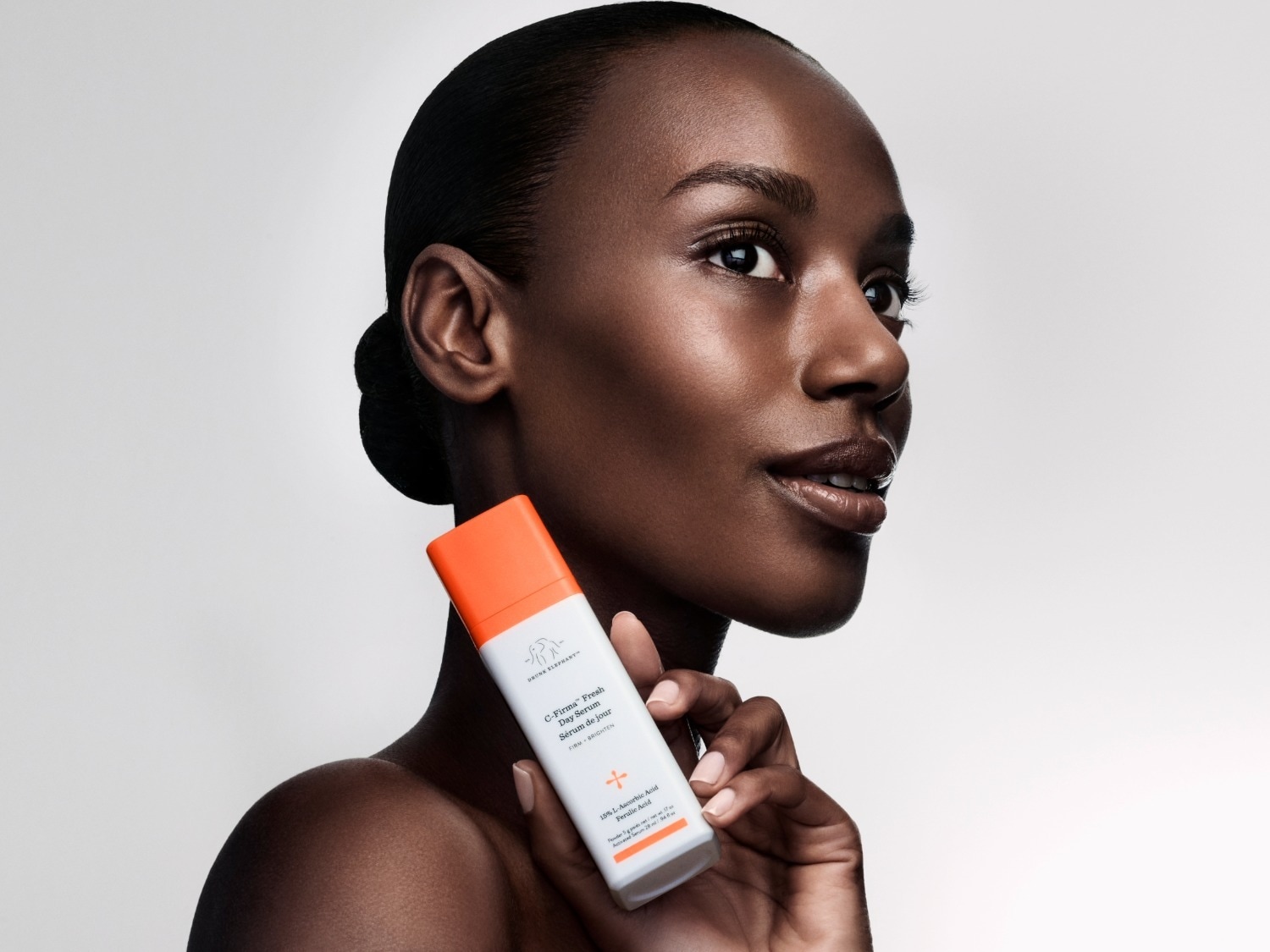Een vrouw met een gladde huid houdt een witte fles Drunk Elephant C-Firma Fresh Day Serum vast, met een oranje dop en de tekst "FIRM + BRIGHTEN" en "15% L-Ascorbic Acid Ferulic Acid" erop.