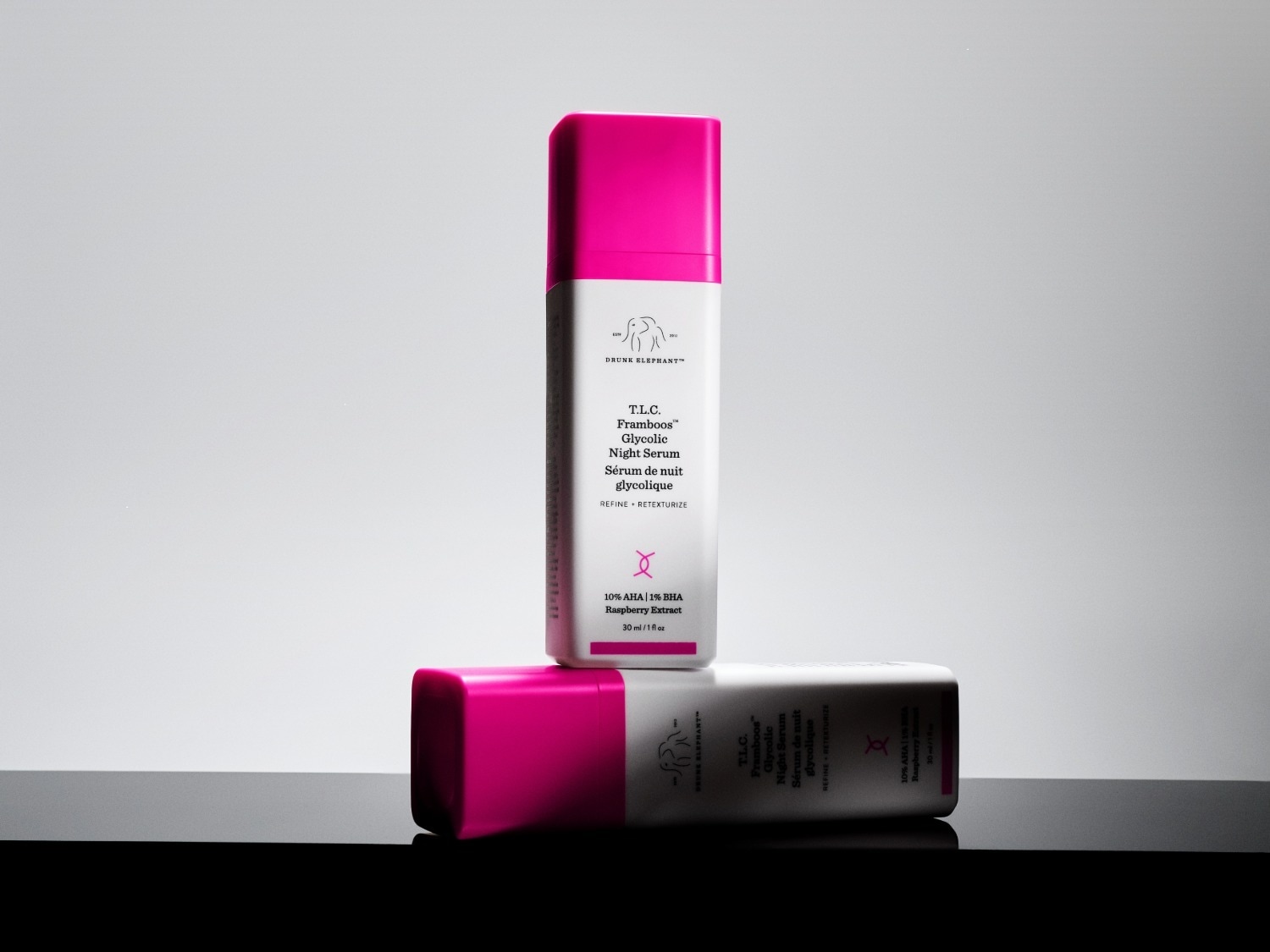 Twee flacons van Drunk Elephant T.L.C. Framboos Glycolic Night Serum, met een roze dop en een witte behuizing, staan op een donker oppervlak tegen een lichte achtergrond.
