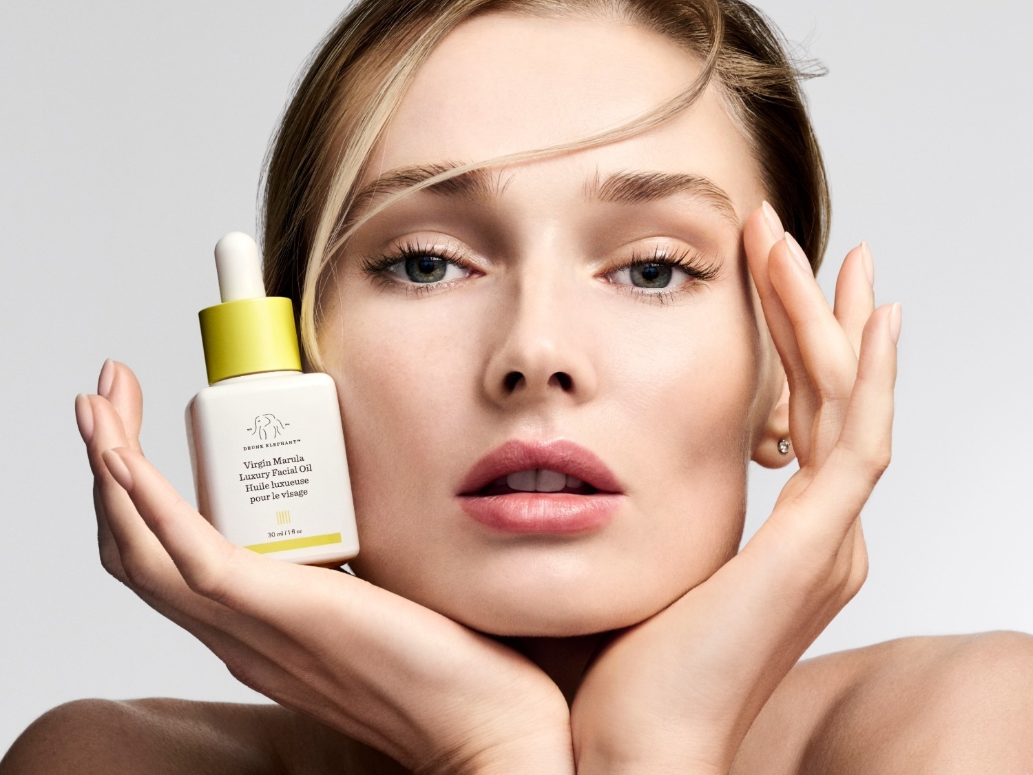 Une femme aux cheveux blonds tient une bouteille blanche de 30 ml d'huile de soin pour le visage Virgin Marula Luxury Facial Oil de Drunk Elephant, avec un bouchon jaune vif et un compte-gouttes blanc, près de son visage.