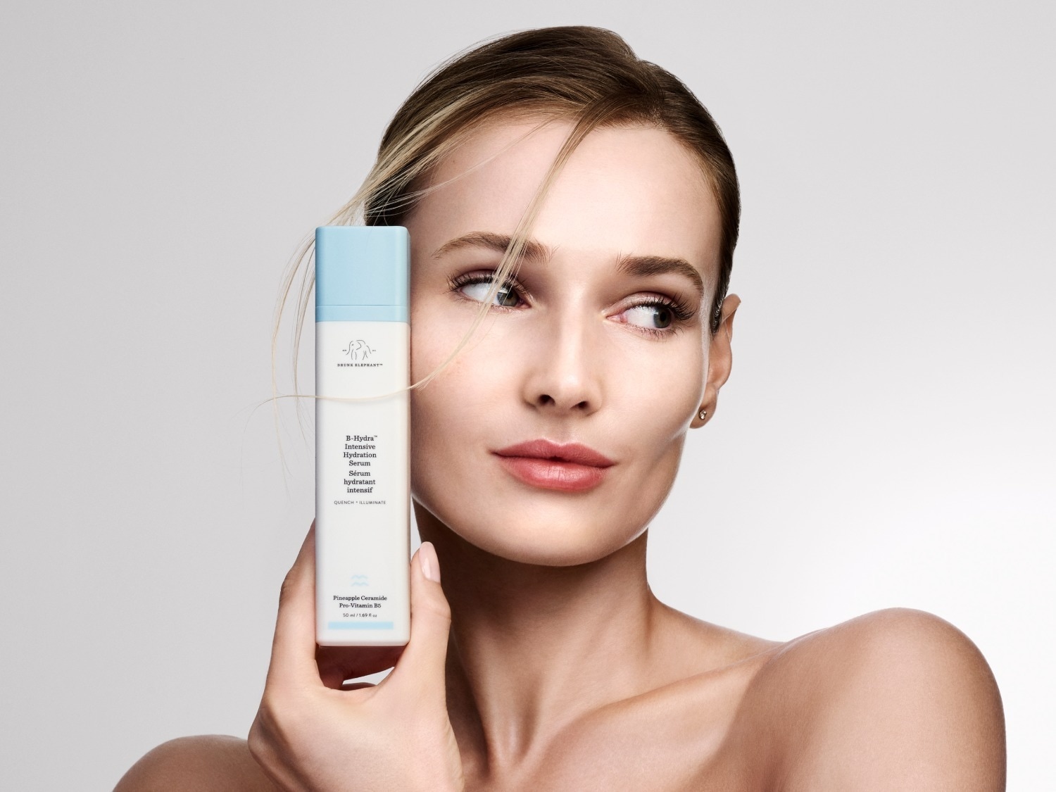 Een vrouw met blond haar en een lichte huidskleur houdt een witte fles Drunk Elephant B-Hydra Intensive Hydration Serum vast, met een lichtblauwe dop en zwarte tekst, terwijl ze naar rechts kijkt.