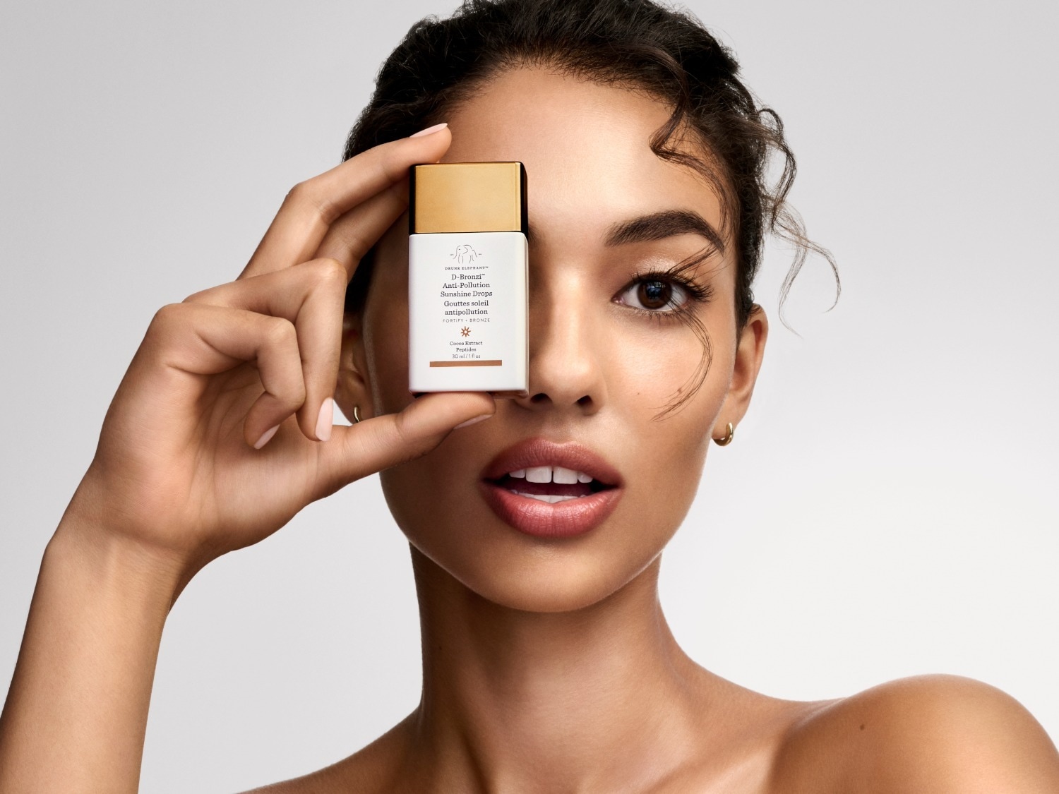 Een vrouw met een gebruinde huid houdt een witte fles Drunk Elephant D-Bronzi Anti-Pollution Sunshine Drops met een gouden dop voor haar linkeroog.