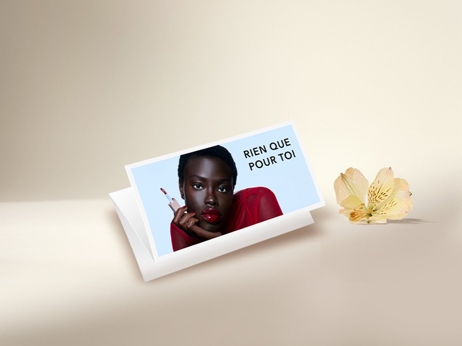 Une carte-cadeau blanche avec une image d'une femme aux lèvres rouges et le texte "RIEN QUE POUR TOI" est posée à côté d'une fleur jaune pâle.