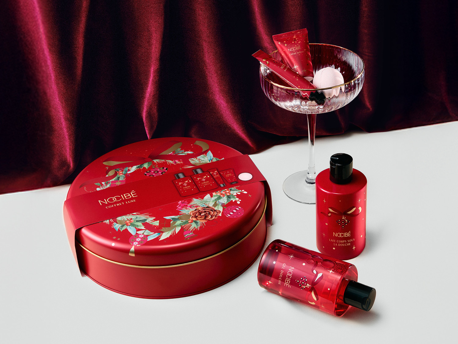 Découvrez le coffret cadeau de luxe Nocibé, un ensemble festif comprenant une boîte ronde rouge ornée de motifs de Noël, deux flacons de gel douche et de lait corporel, ainsi qu'une coupe élégante contenant des crèmes pour les mains et une bombe de bain, le tout sur un fond de tissu rouge velours.