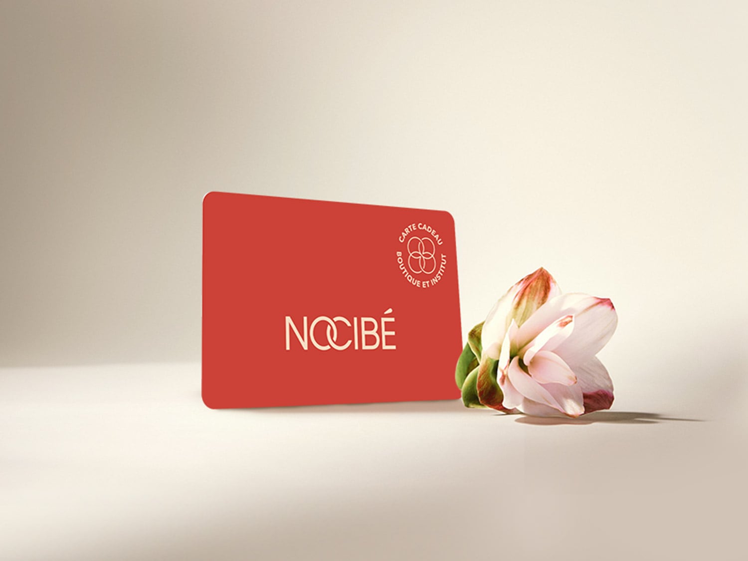 Une carte cadeau rouge vif de Nocibé, avec le logo de la marque en blanc et un cercle indiquant "Carte Cadeau Boutique et Institut", est posée à côté d'une fleur délicate aux pétales blancs et roses, sur un fond clair.