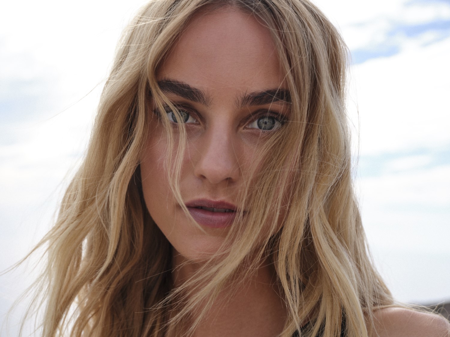 Nahaufnahme einer Frau mit blonden, gewellten Haaren, die ihr Gesicht umrahmen, hellen Augen und einem natürlichen Make-up, das ihre Gesichtszüge betont.