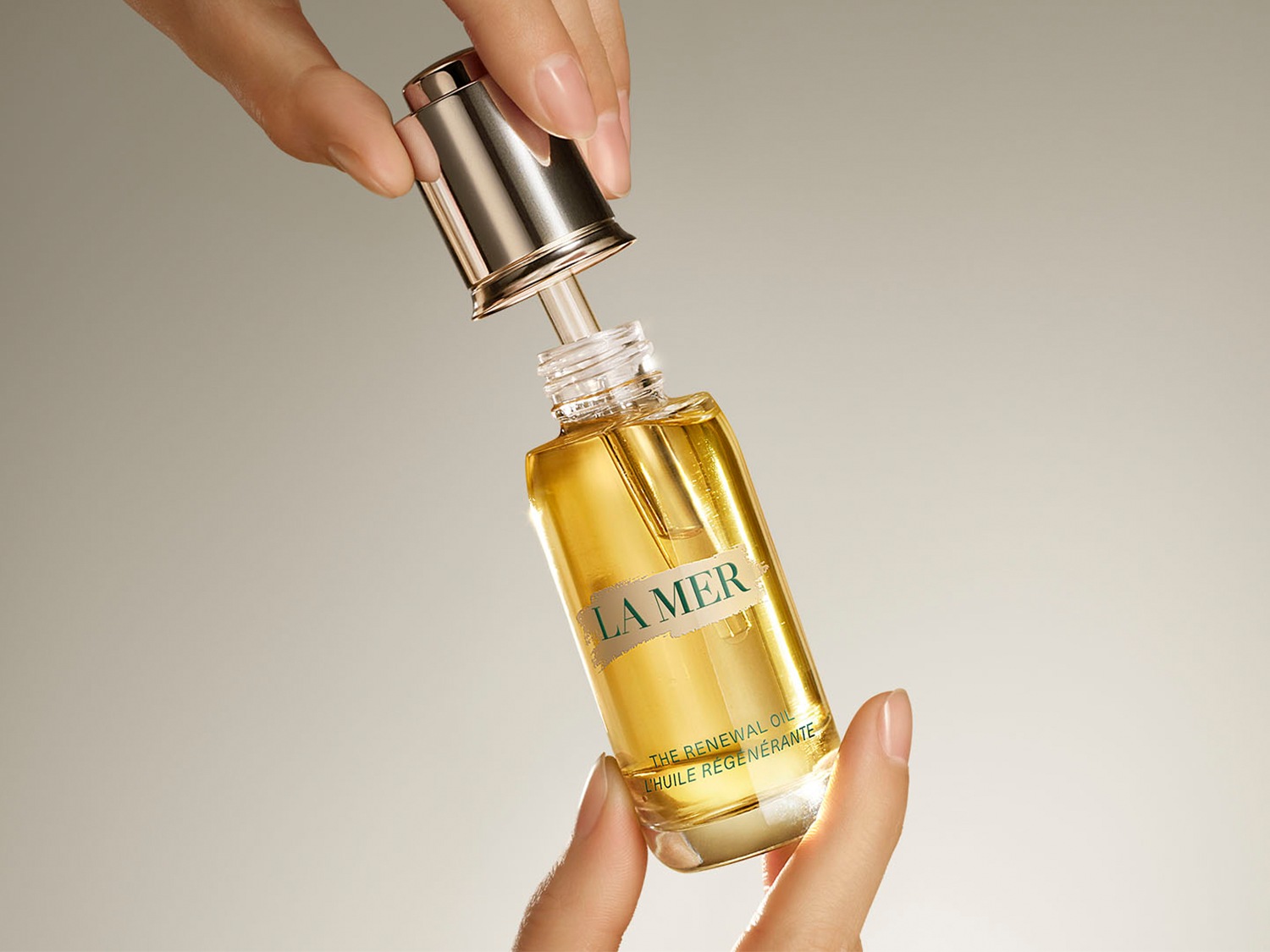 Gouden La Mer Renewal Oil in een heldere glazen fles wordt vastgehouden, met de zilveren dop zwevend erboven, klaar om de huid te vernieuwen.