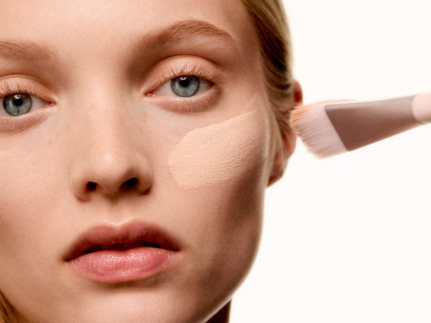 Šviesiaplaukė moteris tepa kreminės spalvos makiažo pagrindą ant veido šepetėliu, kad oda būtų lygi ir nepriekaištinga.