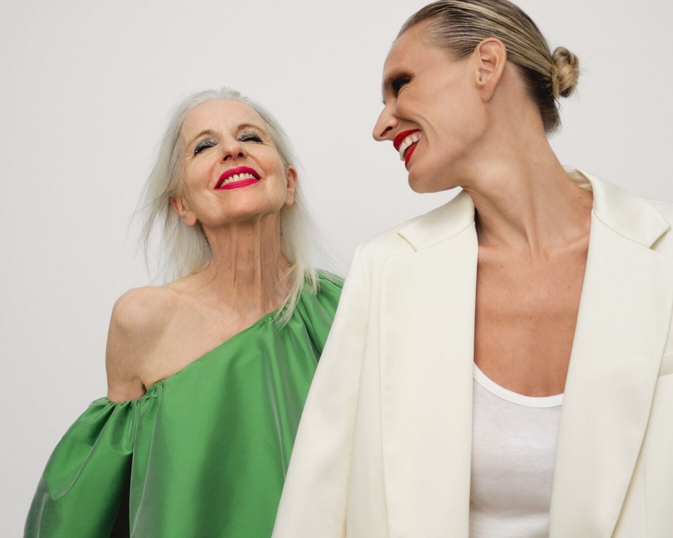 Šviesiame fone matomos dvi moterys: viena su žalia suknele ir ryškiai raudonu lūpdažiu, o kita su šviesiu švarku ir raudonu lūpdažiu.