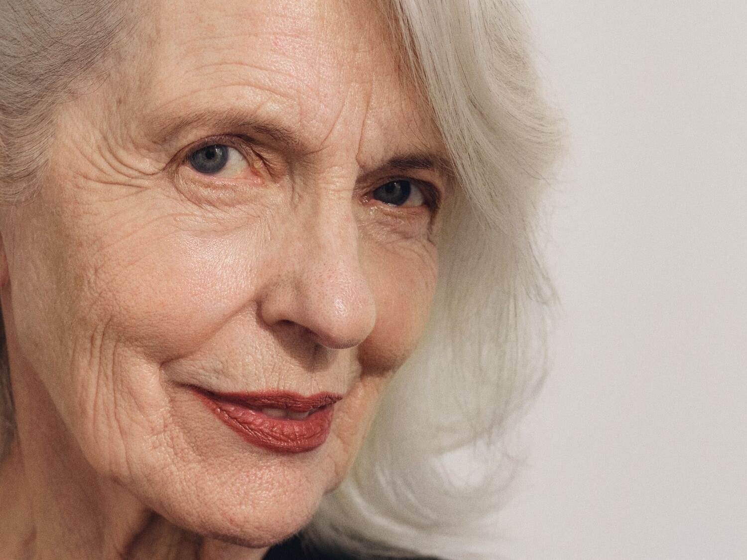 Nahaufnahme einer älteren Frau mit grauen Haaren, blauen Augen und einem sanften Lächeln, das mit einem dezenten, roten Lippenstift betont wird.