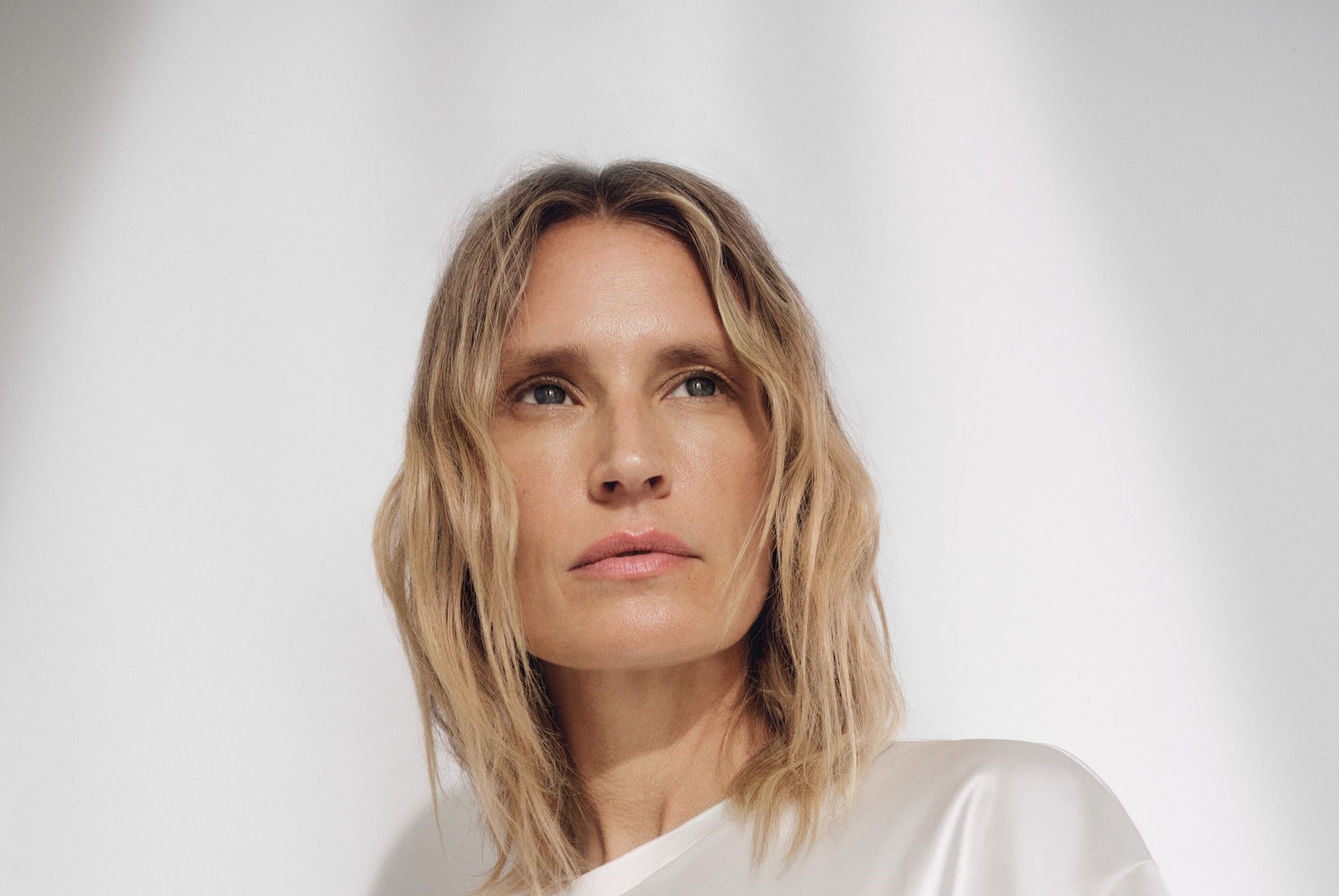 Eine Frau mit blonden, gewellten Haaren und einem hellen Teint blickt mit einem sanften Ausdruck nach oben.