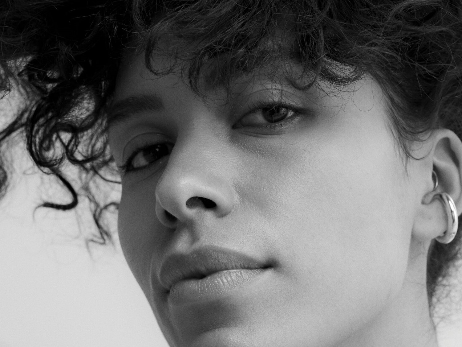 Nahaufnahme einer Frau mit lockigem Haar und silbernem Ohrring, die direkt in die Kamera blickt.