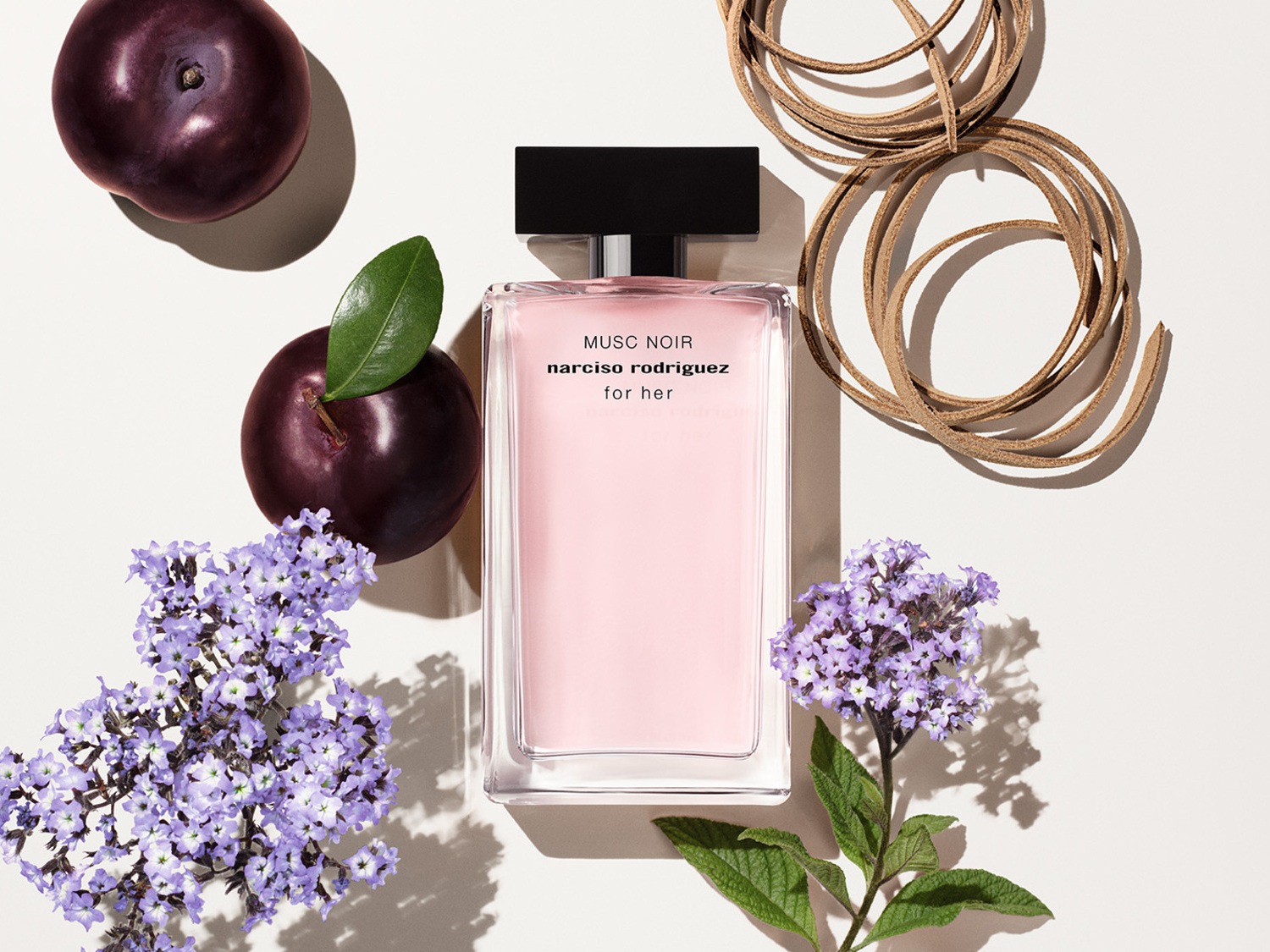 Découvrez le parfum Narciso Rodriguez Musc Noir for Her, présenté dans un flacon rose délicat, entouré de prunes mûres, de fleurs lilas et d'un cordon en cuir brut.