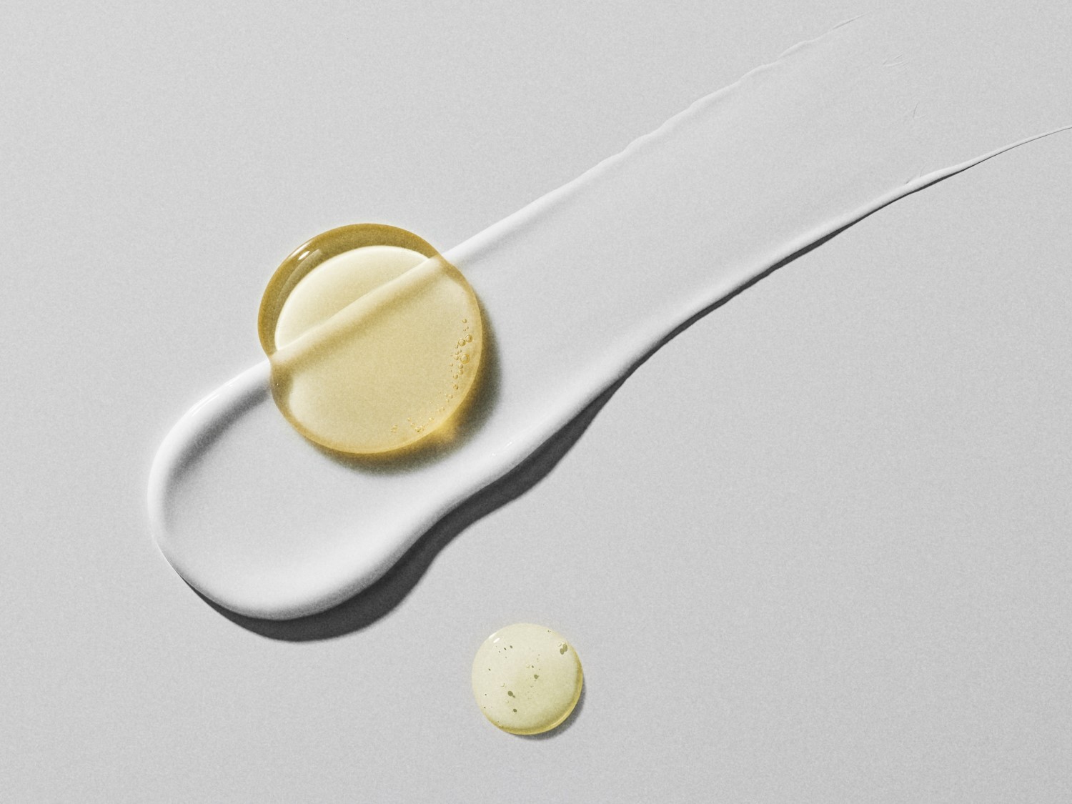 Nahaufnahme eines weißen Cremestrichs mit einem klaren, gelblichen Öltröpfchen darauf und einem kleineren Tröpfchen daneben, das die Textur und Konsistenz der Produkte hervorhebt.