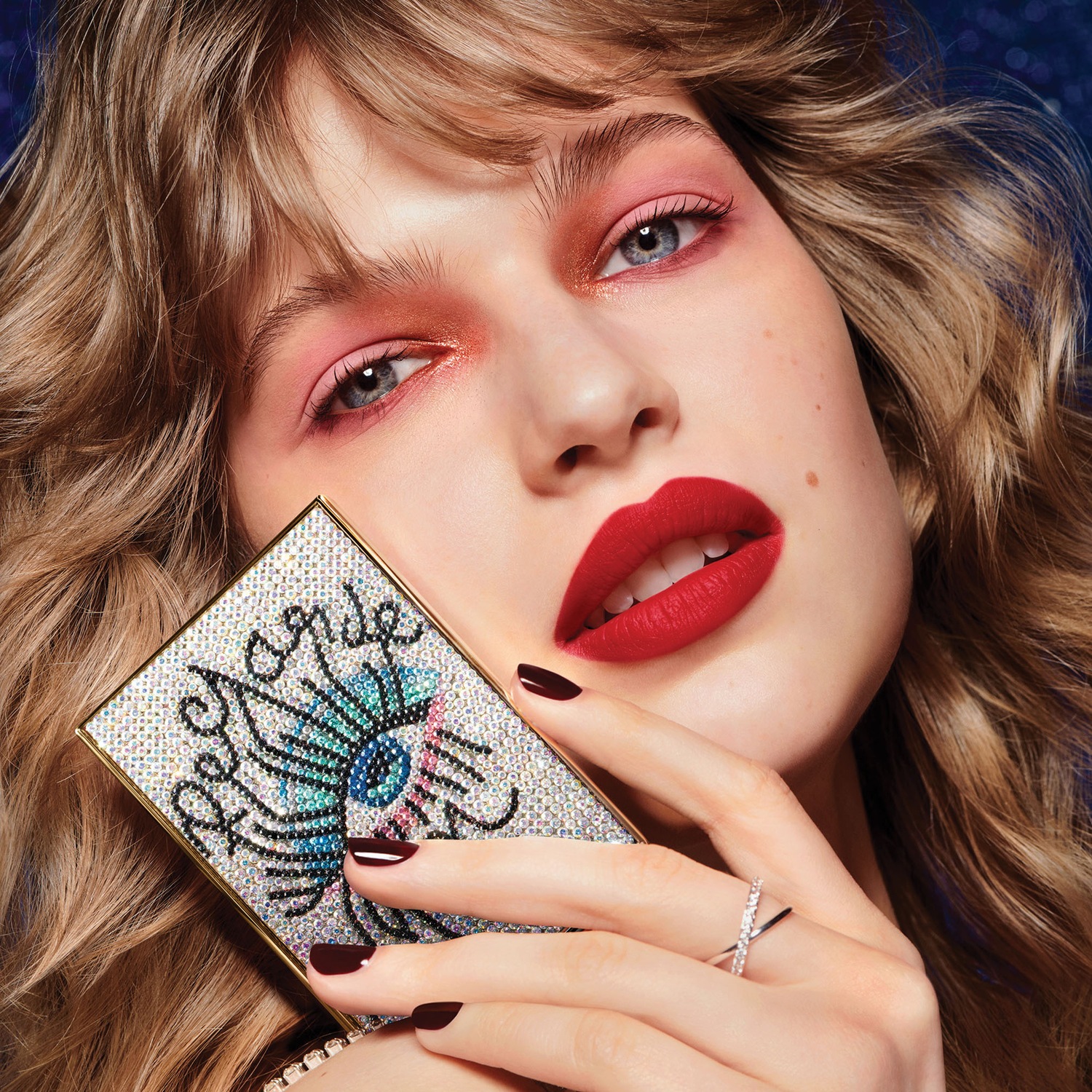 Gros plan d'une femme avec un fard à paupières rose et des lèvres rouge vif, tenant une palette de maquillage ornée de strass.