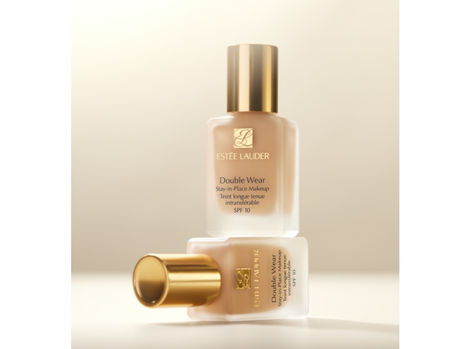 Dwa flakony podkładu Estée Lauder Double Wear Stay-in-Place Makeup SPF 10, jeden na drugim, z złotymi zakrętkami, na beżowym tle.